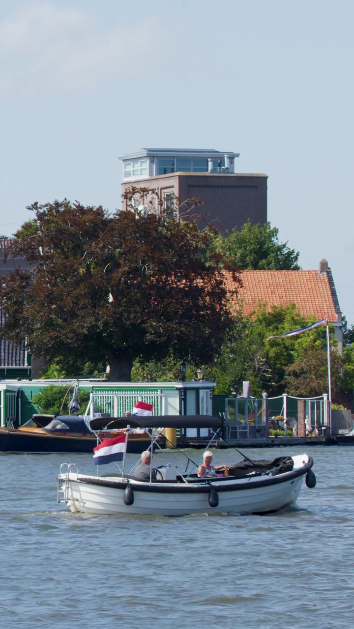 Una piccola barca a motore con passeggeri si muove lungo un fiume a Zaandam, passando accanto a case tradizionali olandesi sotto la luce del giorno intensa, con un'inquadratura stabile della telecamera