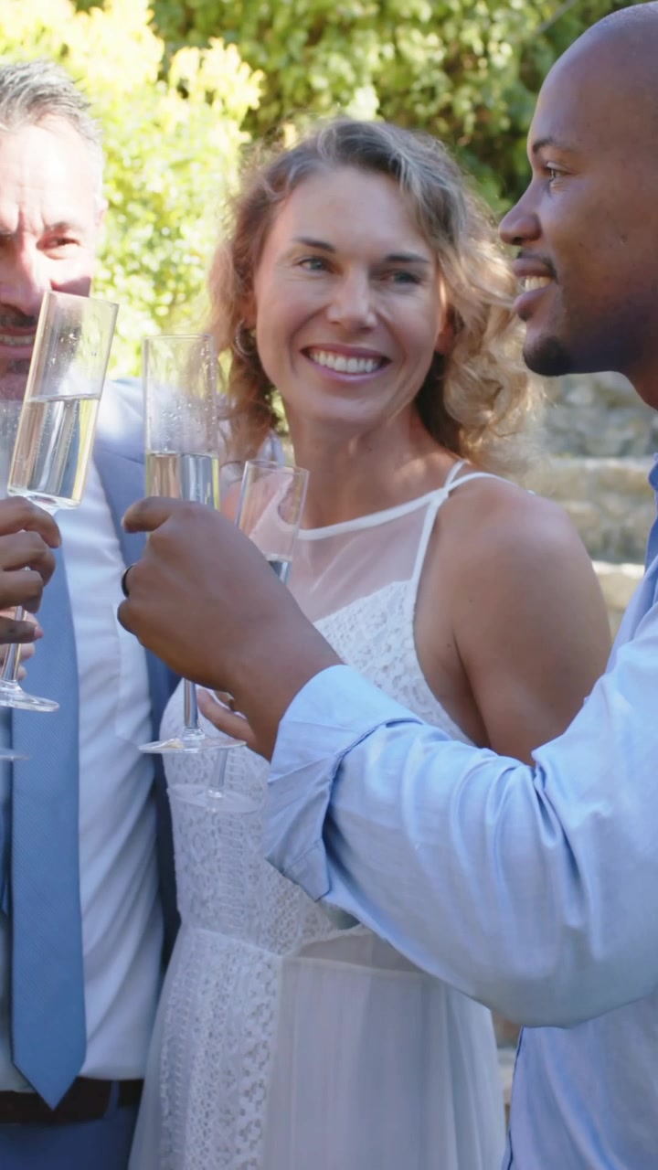 Vertical video: Raising bride in white dress with men on sunny steps, clinking flutes, showing ring