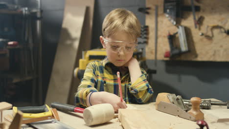 niño pequeño y lindo caucásico con gafas trabajando en un taller de carpintería y dibujando con lápiz