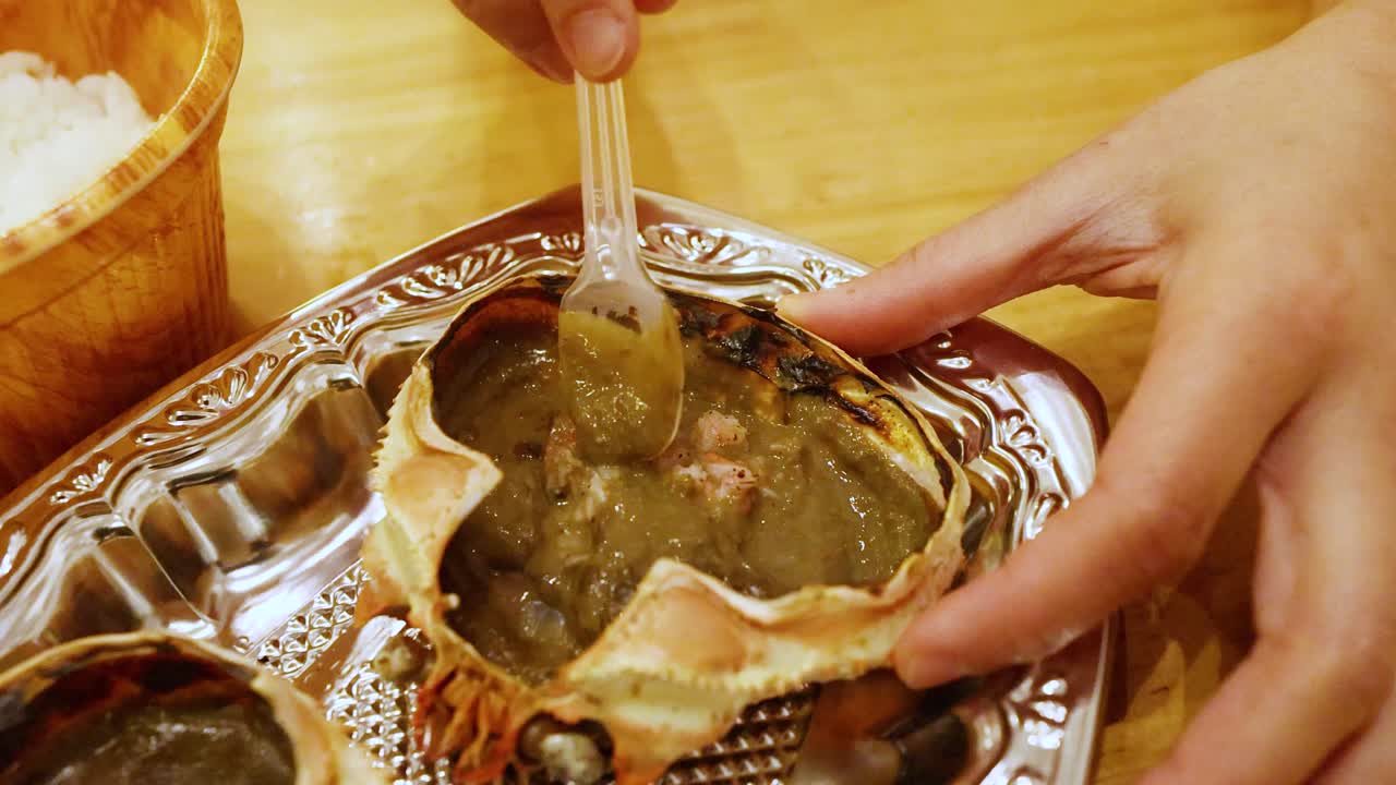 Primo piano di mani che usano un cucchiaio di plastica per mangiare miso di granchio grigliato direttamente dal guscio.