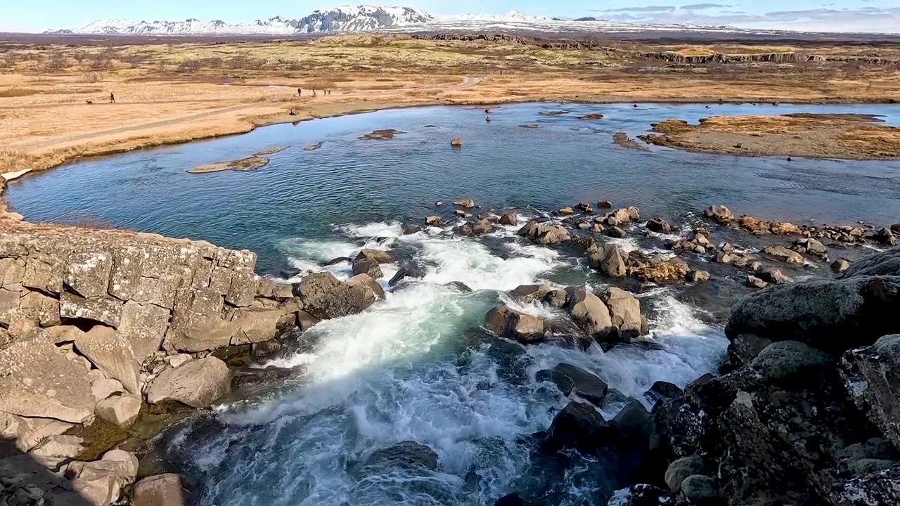 Uma fotografia em grande angular de um rio azul claro fluindo sobre rochas na natureza selvagem islandesa, com montanhas distantes cobertas de neve e uma vasta paisagem de tundra.