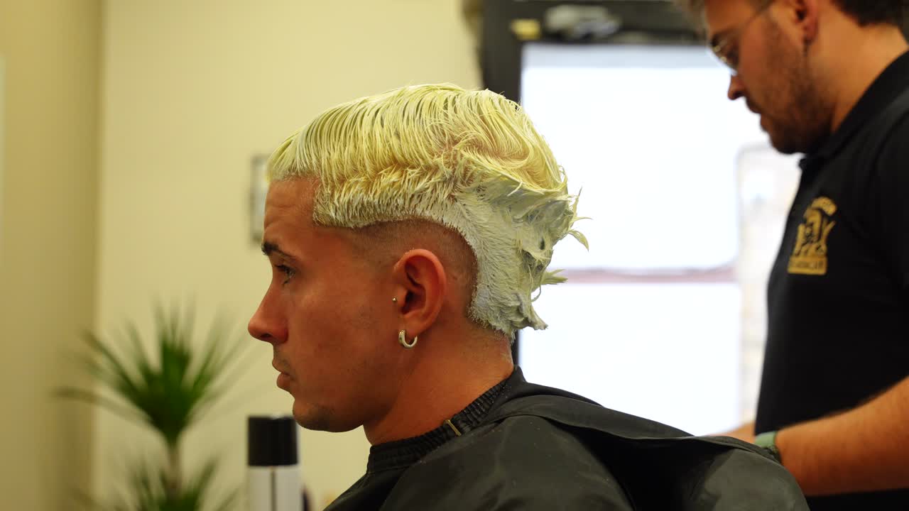 Close up of a man with wet yellow hair while hairdresser scoops bleaching mass with spatula to continue application