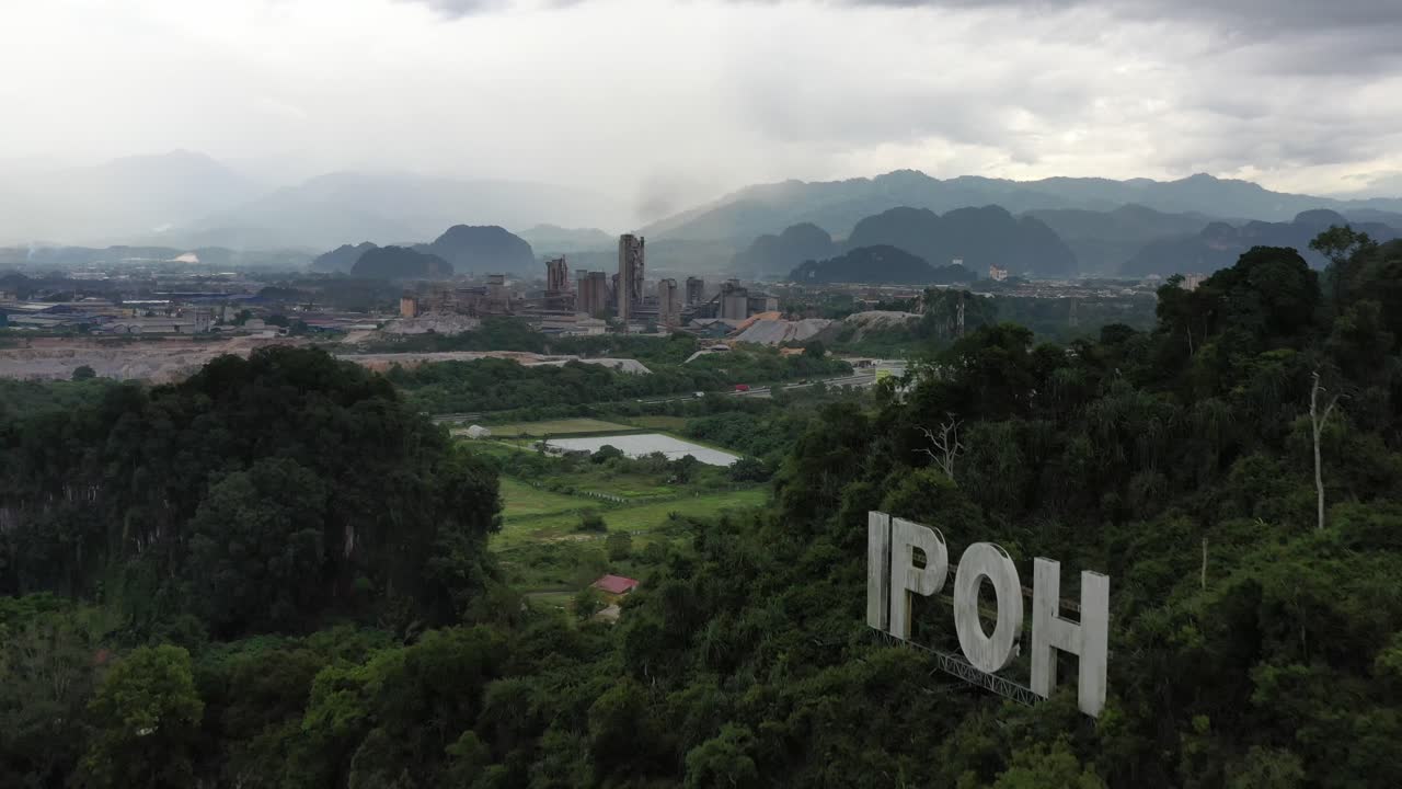 Drone flyover limestone hill with ipoh sign on top, overlooking at kawasan perindustrian tasek with excavation and active quarrying operation site and beautiful natural landscape in the background.