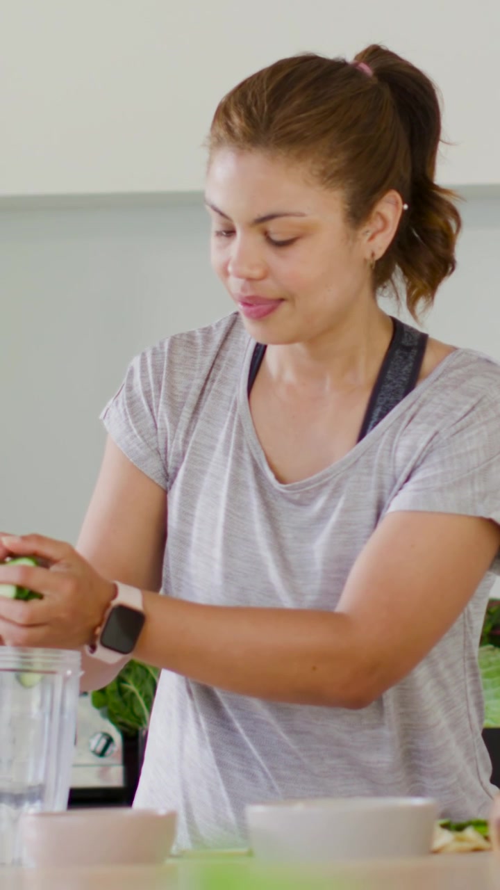 Video verticale: Donna in maglietta che lancia un'occhiata, strappa verdure a foglia e carica il frullatore in cucina per un succo, con un orologio