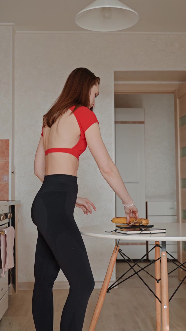White woman at home walking to table placing sandwich and drink on dining table wearing red sports top and black leggings, casual postworkout snack break, minimalist apartment interior with natural