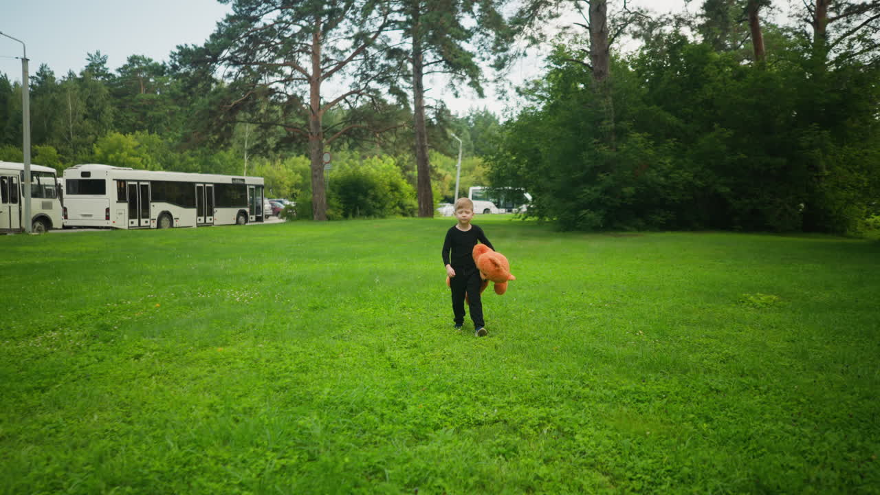 Front view of young boy dressed in black outfit carrying orange teddy bear while walking on large green field surrounded by trees and parked buses in peaceful park like environment under open sky