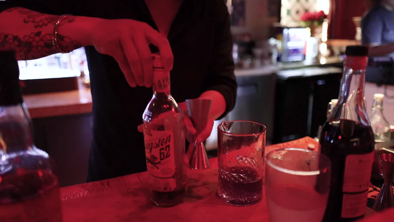 Bartender pours clear liquor into jigger, preparing cocktail at bar with moody red lighting