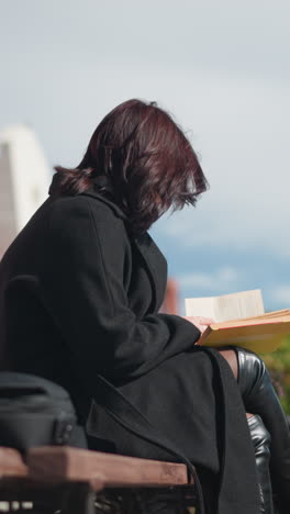joven con abrigo negro leyendo su libro al aire libre, centrándose en las páginas mientras su cabello sopla en el viento, edificios urbanos, postes de lámparas y personas que pasan forman un fondo suave y borroso
