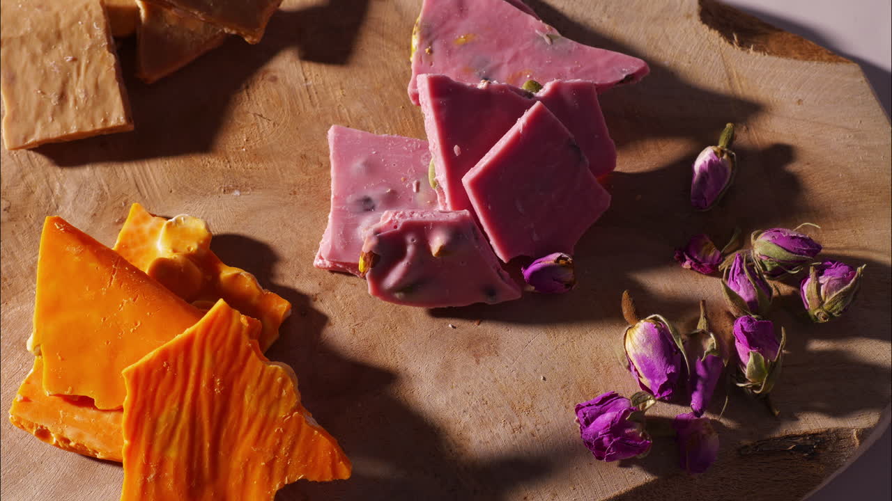 A hand reaches for a vibrant orange-colored brittle piece on a wooden board, placed beside pink strawberry chocolate chunks and scattered dried rosebuds, in soft shadow and natural kitchen light.