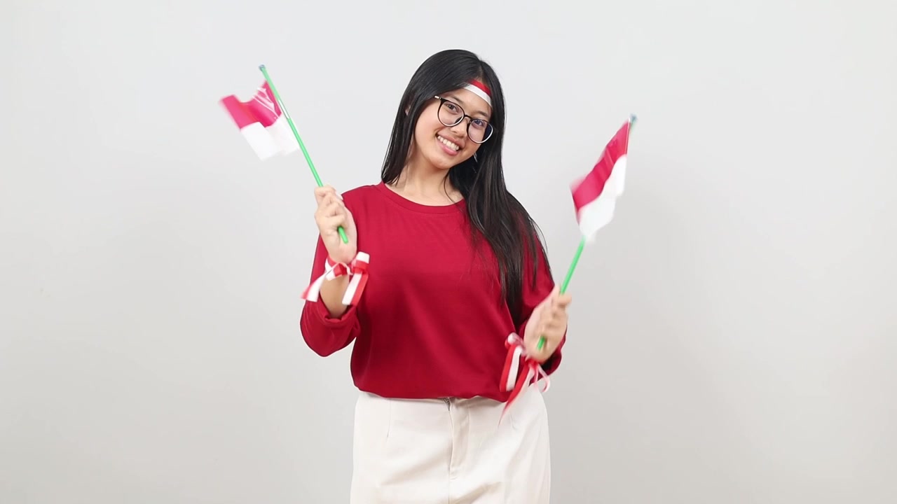 Una donna indonesiana con un nastro rosso e bianco, che tiene e sventola una bandiera rossa e bianca, sorride felicemente alla macchina fotografica, in piedi isolata su uno sfondo bianco.