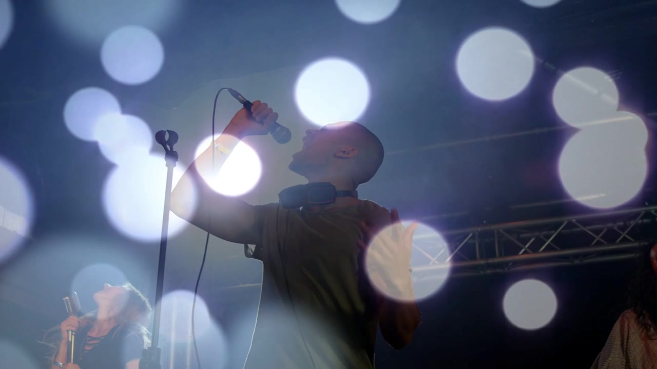 Music lead singer leaning, singing into mic, backup singers moving, bokeh overlay obscuring singers