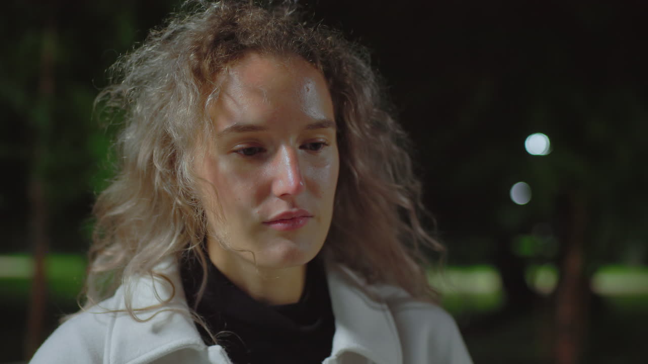 Girl seated outdoors at night slowly lifting head in quiet reflection with curly hair gently swaying in night breeze, surrounded by blurred background of trees and soft glowing park lights
