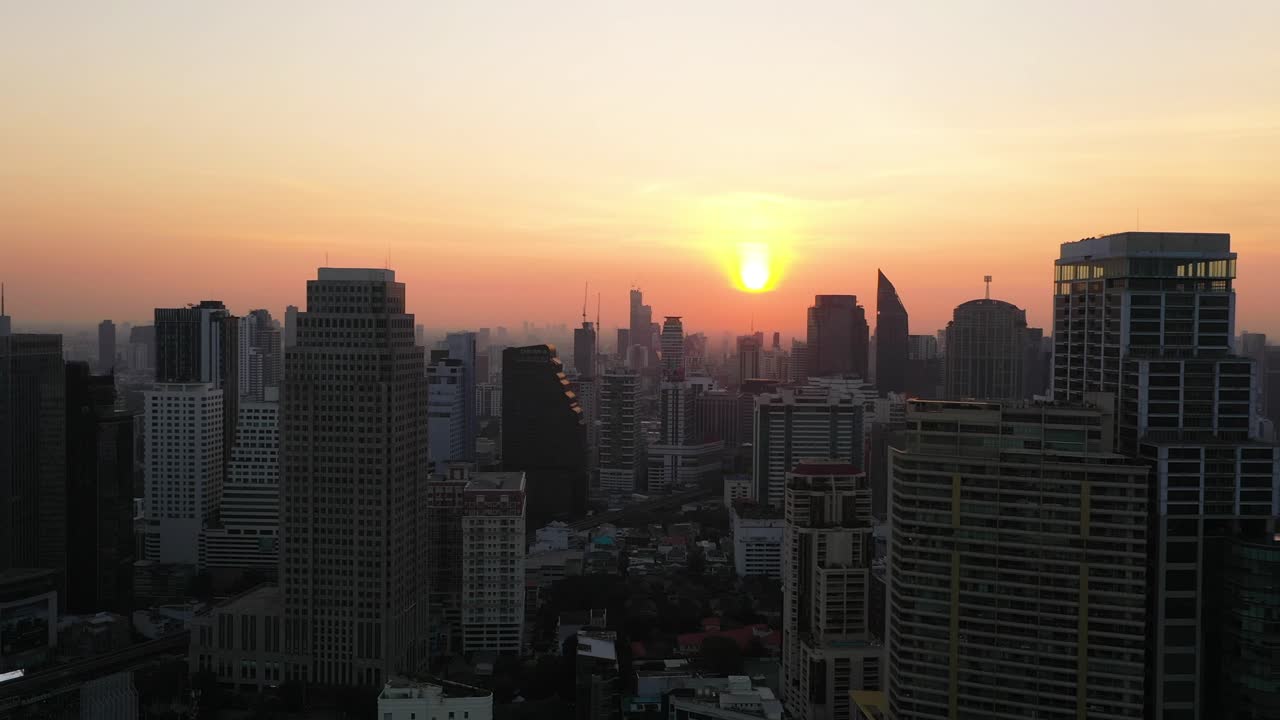 Aerial flying sideways alongside buildings, skyline and skyscrapers of Sukhumvit district during colorful red sunrise in Bangkok, Thailand.