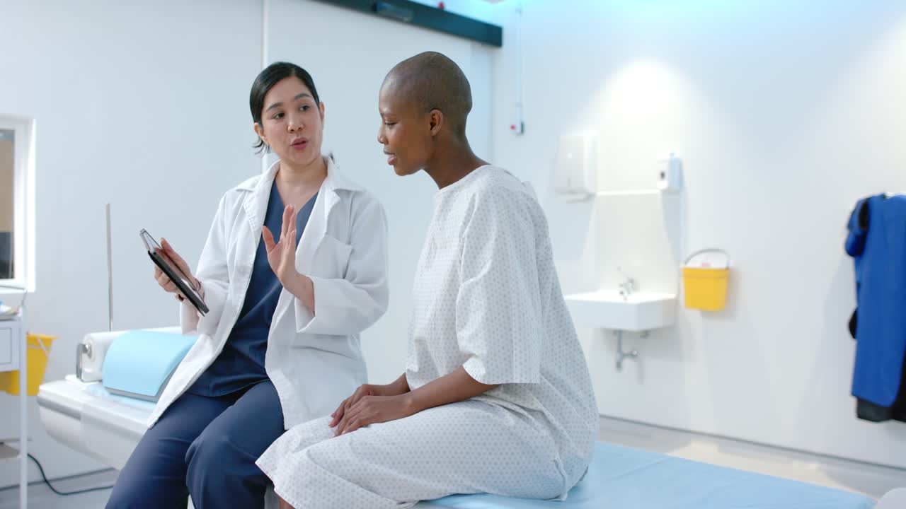 Medico asiatico in camice tocca il tablet, mostrando le cure alla paziente afroamericana sul lettino da visita