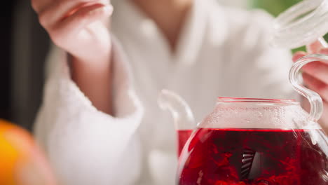 woman stirs hibiscus tea in teapot closeup. lady brews red herbal drink for breakfast at table in morning. homemade healthy organic beverage