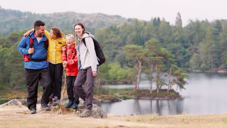 young family on the left of shot standing on a rock by a lake in the countryside looking to camera laughing, lake district, uk