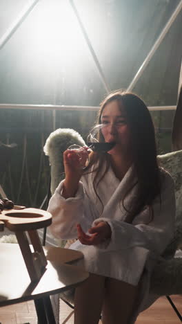 lovers enjoy wine with snacks at round house. couple in terry bathrobes sits in cozy armchairs on glass terrace. young lady trying out grape beverage