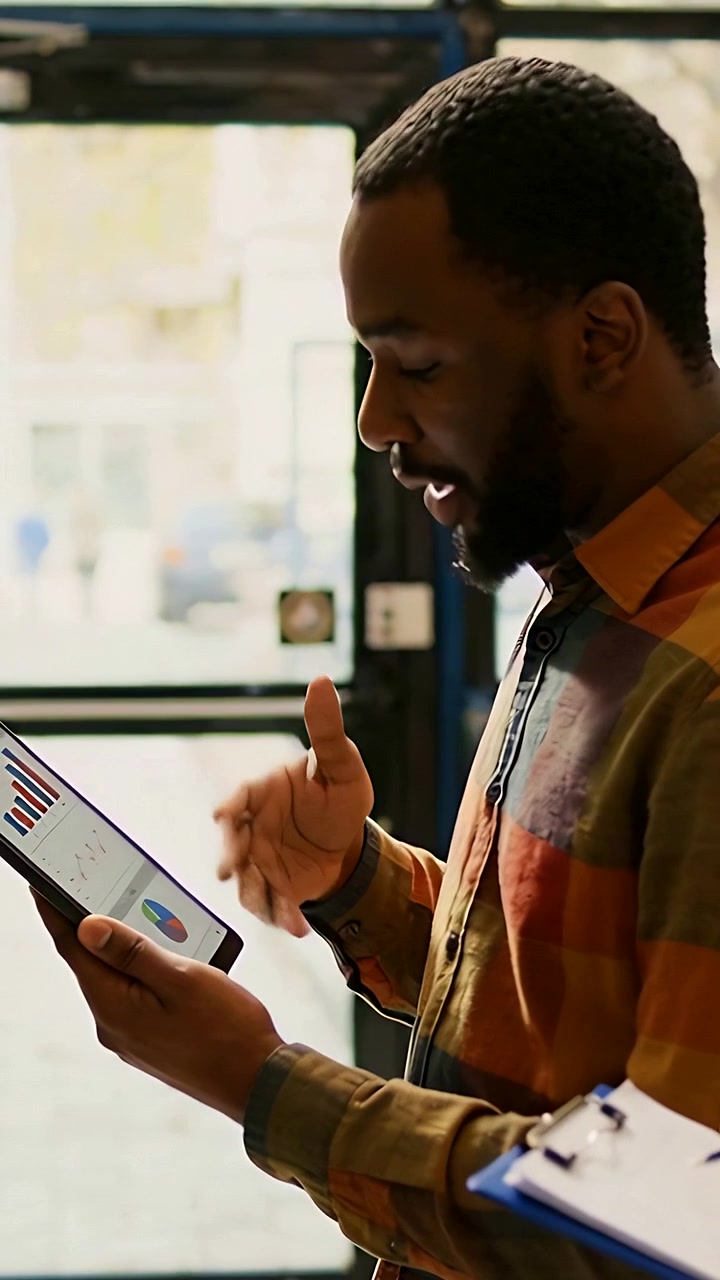 Vertical video: Checking tablet data, man in plaid as coworker holding clipboard by door