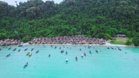 drone aéreo tiró sobre barcos de cola larga en un pueblo de cabañas de madera a lo largo de una playa tropical en tailandia con ladera verde en el fondo