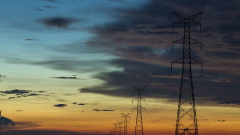 Strommasten-Ragen-Während-Eines-Filmischen,-Farbenfrohen-Blau-orangefarbenen-Sonnenuntergangs-Mit-Sich-Langsam-Bewegenden,-Launischen-Wolken-Bis-Zum-Horizont