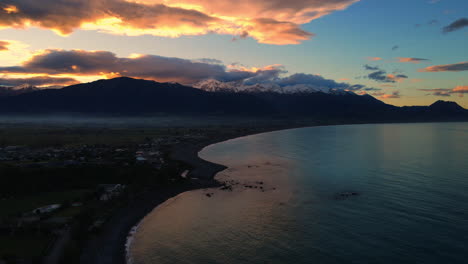 Tiro-De-Dron-Del-Mar-Con-Olas-Rompiendo-Y-Casas-De-Fondo-De-Montaña-Cubiertas-De-Nieve-Blanca-Naranja-Atardecer-Amanecer-Cielo-Nueva-Zelanda