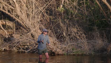 male fly fisherman in hat and sunglasses casts his fishing rod while knee deep in water during golden hour in slow motion