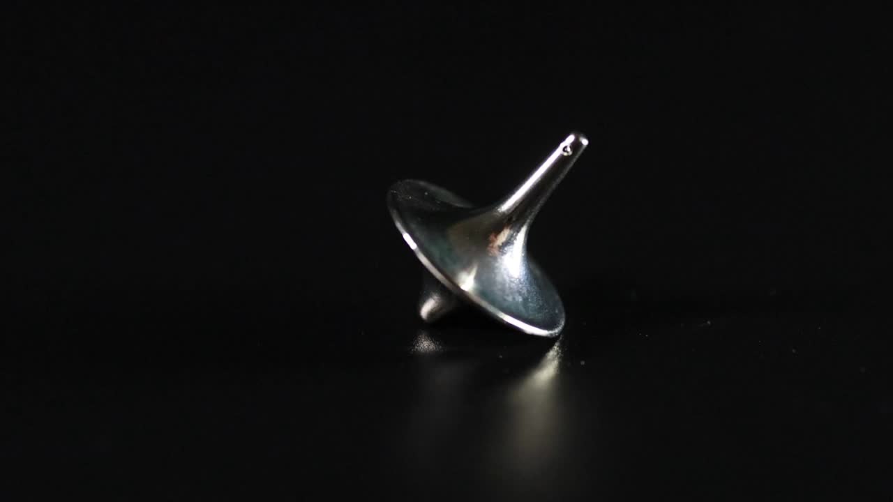 Fingre snurrer en sølvfarvet metal snurretop på en sort, reflekterende overflade under dramatisk belysning