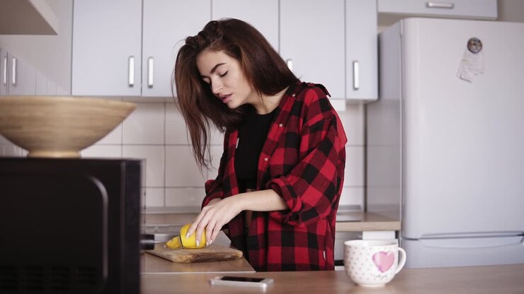 Young beautiful girl in red flannel shirt slicing lemon and then making herself a cup of tea in the kitchen.
