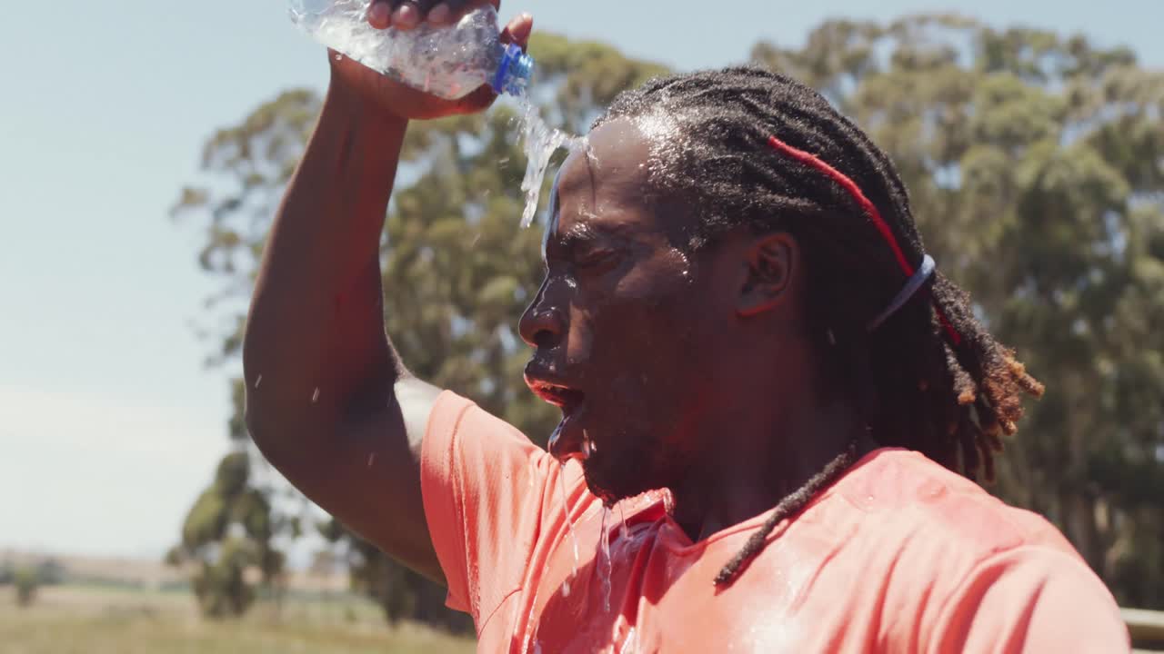 Un homme afro-américain sportif avec des dreadlocks se versant de l'eau sur la tête après l'exercice sous le soleil.
