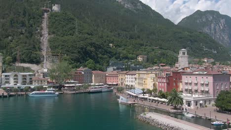 Drohne-Fliegt-Auf-Den-Platz-Im-Zentrum-Der-Norditalienischen-Stadt-Riva-Del-Garda-Zu,-Mit-Dem-Gardasee-Im-Vordergrund-Und-Den-Alpen-Im-Hintergrund