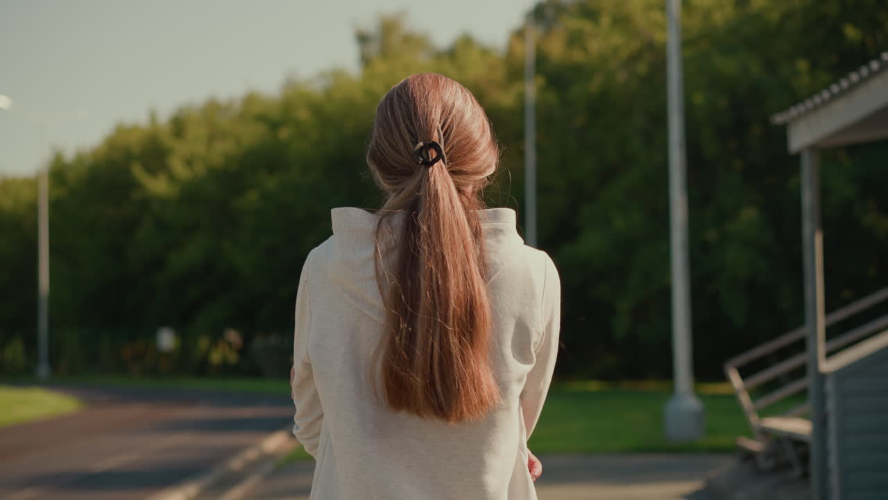 후드 스웨터를 입고 머리카락을 뒤로 고 은 날 빈 경기장 트랙을 따라 걸어가는 여성의 뒷면, 따뜻한 빛이 그녀를 비추는 동안 생각깊게 팔을 습니다.