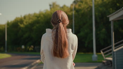 back view of a woman in a hooded sweatshirt with her hair tied back, walking along an empty stadium track on a sunny day, arms folded thoughtfully, as the warm sunlight illuminates her