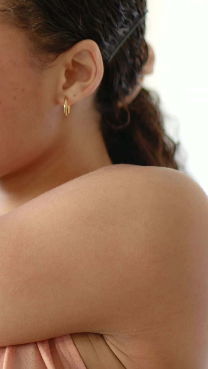 Video verticale: Donna che si tiene il braccio sinistro e si sistema i capelli nella stanza, con orecchino a cerchio dorato e spallina color pesca.