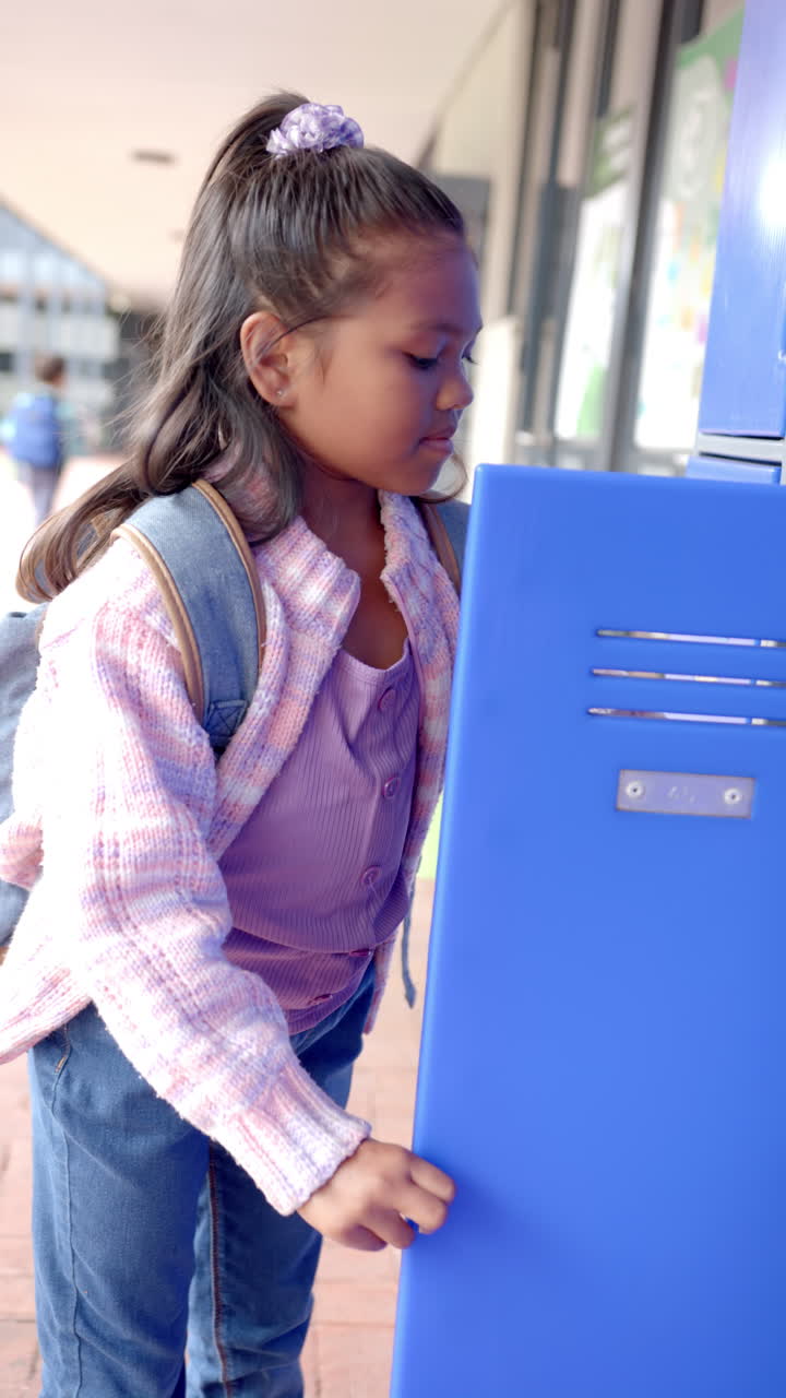 Vertical video: In school, young biracial girl with light brown skin is opening a blue locker