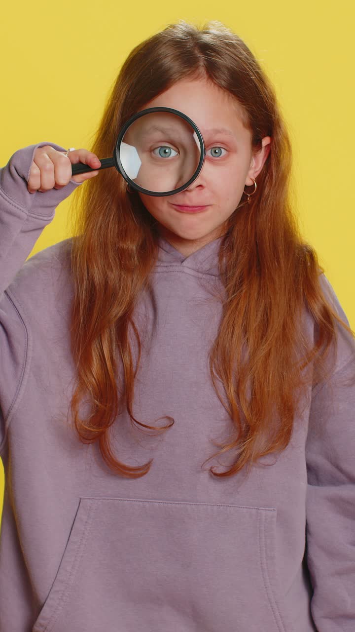 Child girl kid holds magnifying glass near face looking at camera with big zoomed eye analyzing