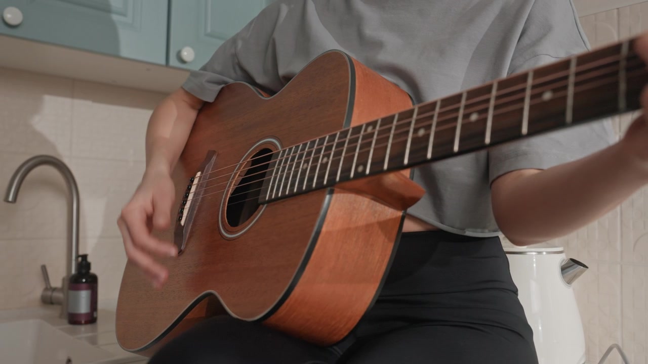Persona che strimpella la chitarra acustica in cucina. Un amante della musica si esercita con gli accordi, primo piano sulle mani, toni caldi del legno, camicia grigia casual, seduto vicino al lavello, una sessione casalinga intima focalizzata sulla tecnica delle dita.
