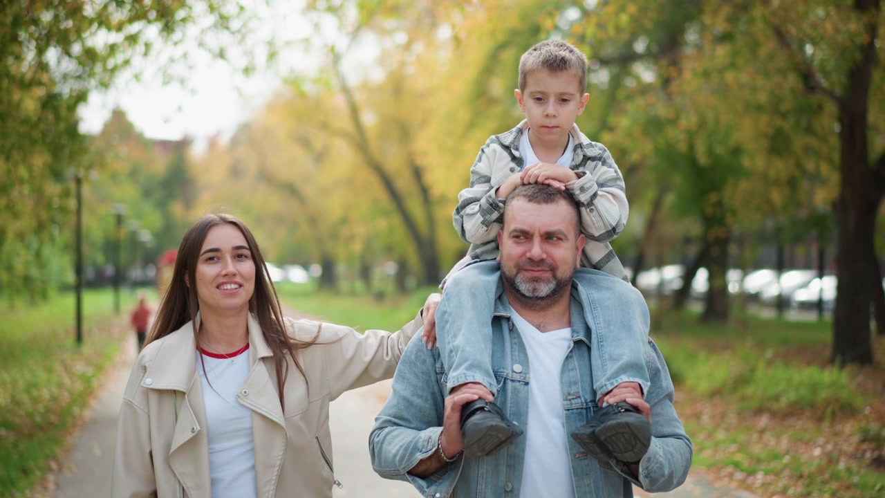 Famiglia che cammina insieme nel parco con un bambino seduto sulle spalle del padre e la madre accanto a loro. La scena presenta una luce naturale soffusa, alberi verdi che fiancheggiano il sentiero e persone che si godono una passeggiata all'aperto.
