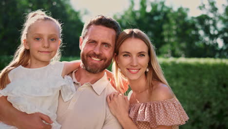 Family-looking-at-camera.-Mother-and-daughter-hugging-father-on-backyard