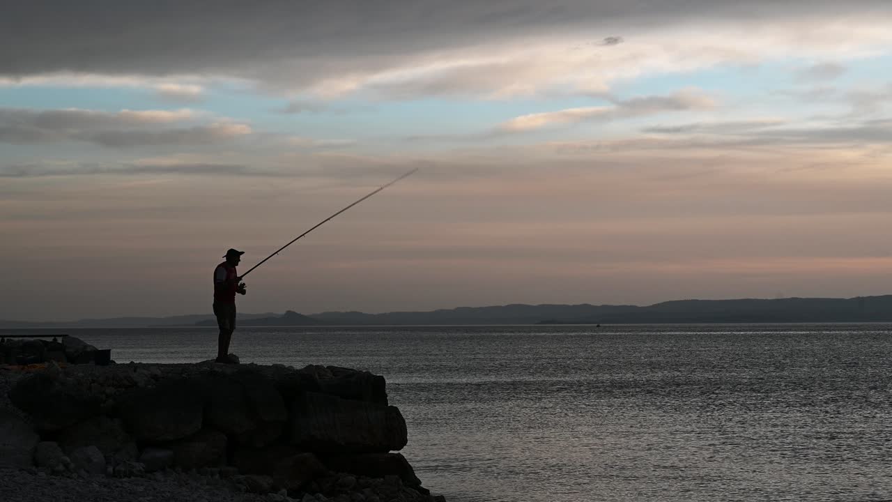 Pria tua memancing dari dermaga batu di danau saat matahari terbenam musim panas yang indah.