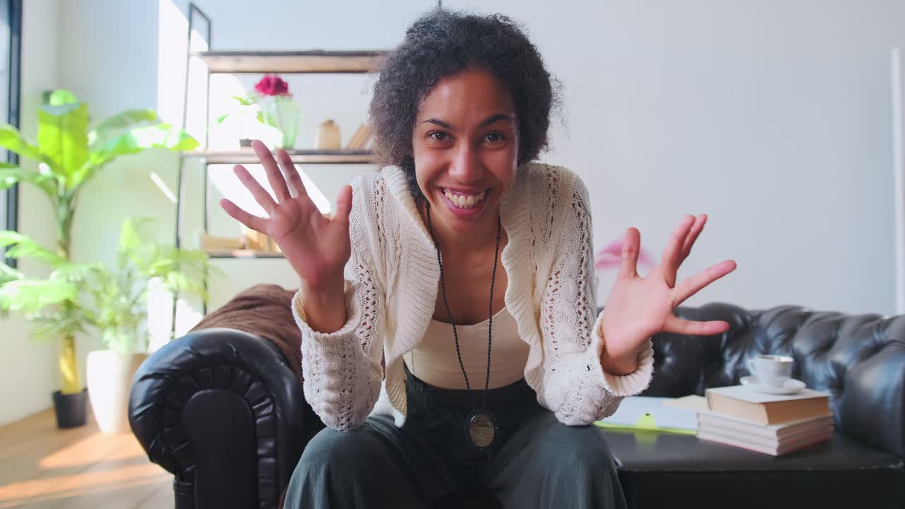 joven mujer afroamericana agita la mano en saludo y sonríe se sienta en el sofá