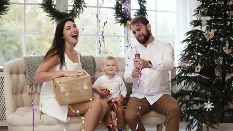 young mother is holding a present in her hands while young father is blowing up a firework, sparkles and confetti flying in the