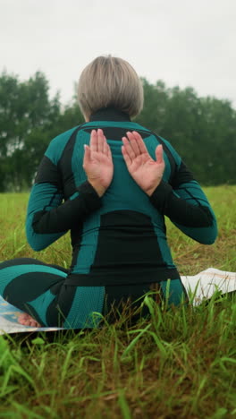 vista trasera de una mujer de mediana edad sentada en una alfombra en la postura de sukhasana practicando yoga con los ojos cerrados, abrazando la calma en un vasto campo de hierba bajo un cielo nublado, con árboles alineados en el fondo