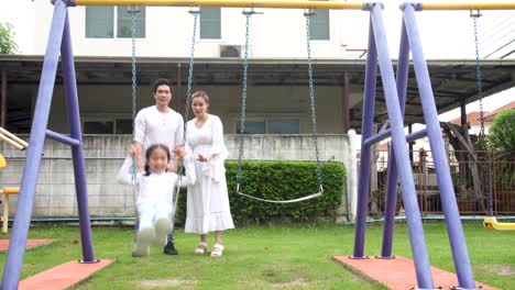 happy family time, parents with daughter playing on a swing in playground, parents pushing little girl, beautiful family is having fun outside.