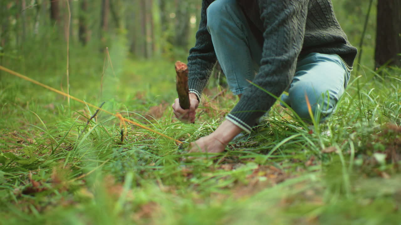 Young man crouches on green forest ground using stick to peg yellow rope while setting up tent during calm camping activity in quiet woodland surrounded by tall trees and natural light