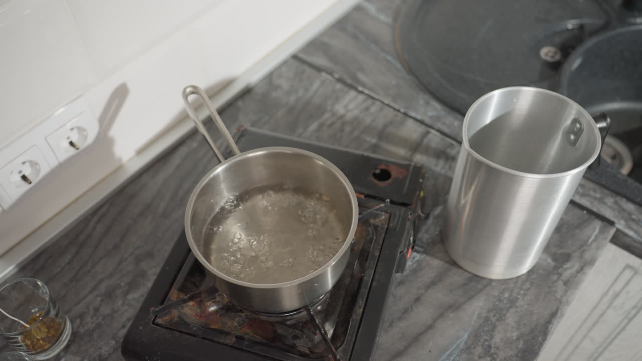 Overhead view of water boiling and steaming vigorously inside stainless steel pot placed on gas burner, with stainless jug and glass cups visible on marble kitchen countertop