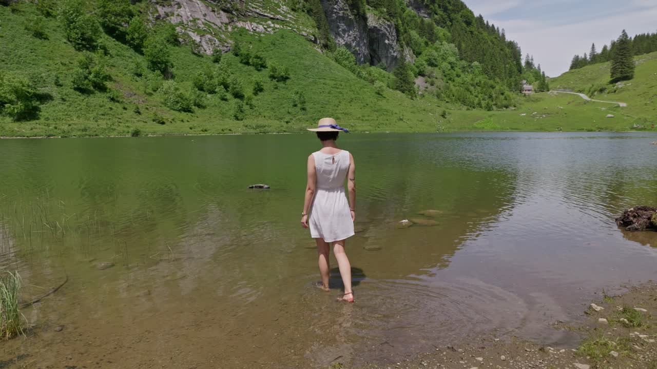 Woman in white dress and hat stands by scenic Talalpsee Lake surrounded by lush green mountains
