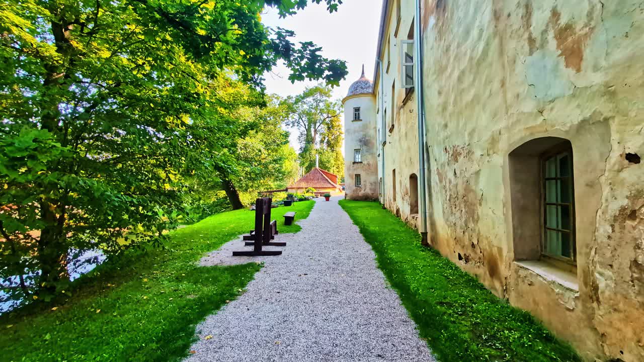 POV shot walking along a gravel path with green grass and a weathered castle wall in Jaunpils, Latvia