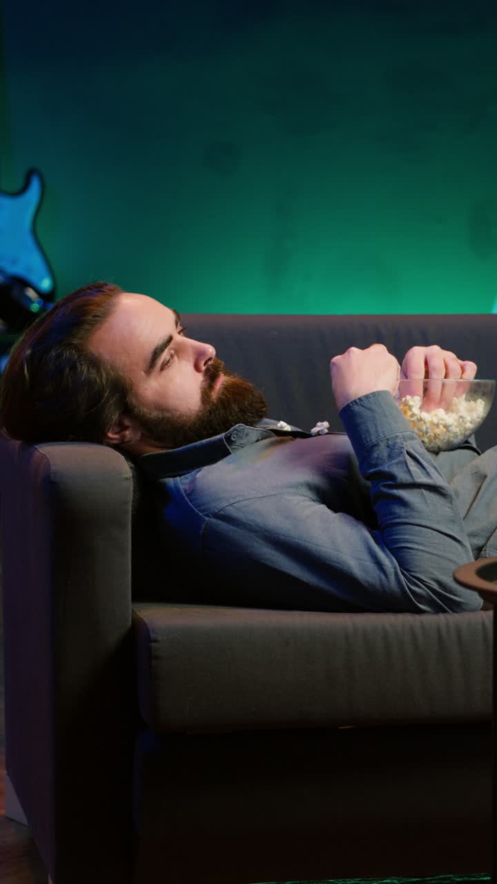Vertical video Bored man procrastinating at home, laying down on couch, watching TV