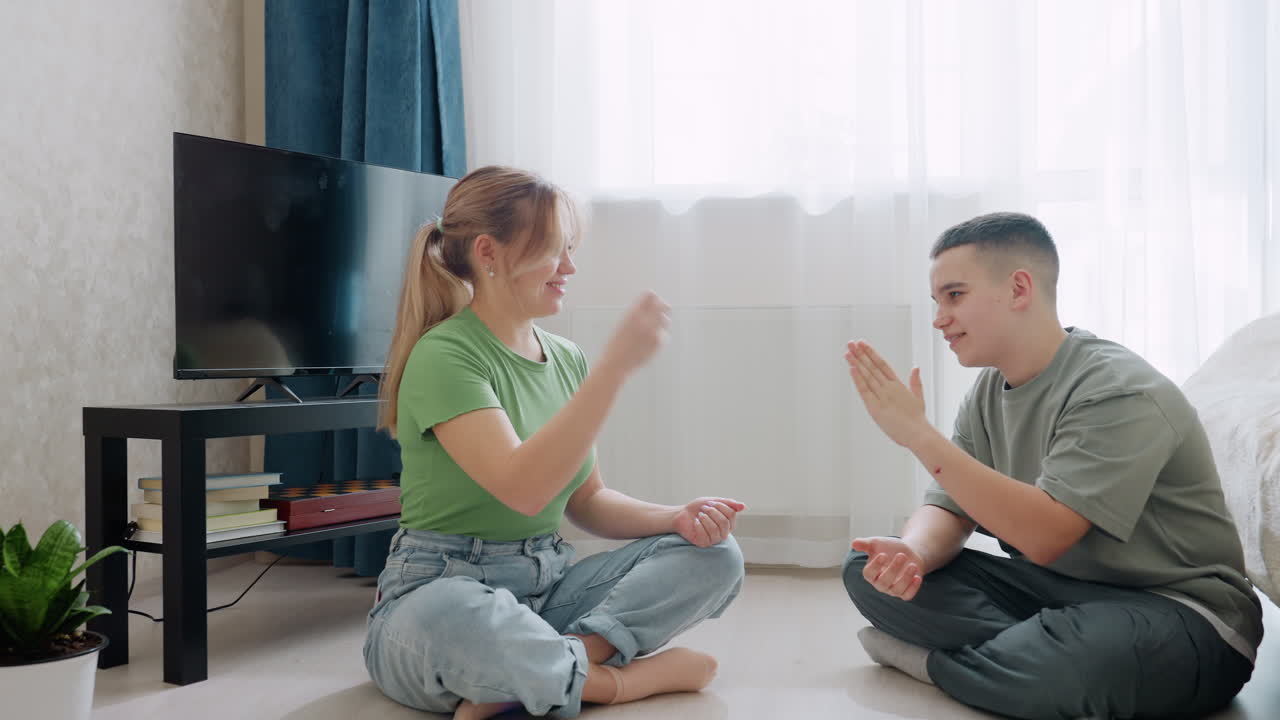 Mother and son sit cross-legged on floor near bed and television playing hand game in bright room, expressing joy, bonding, fun interaction and family connection