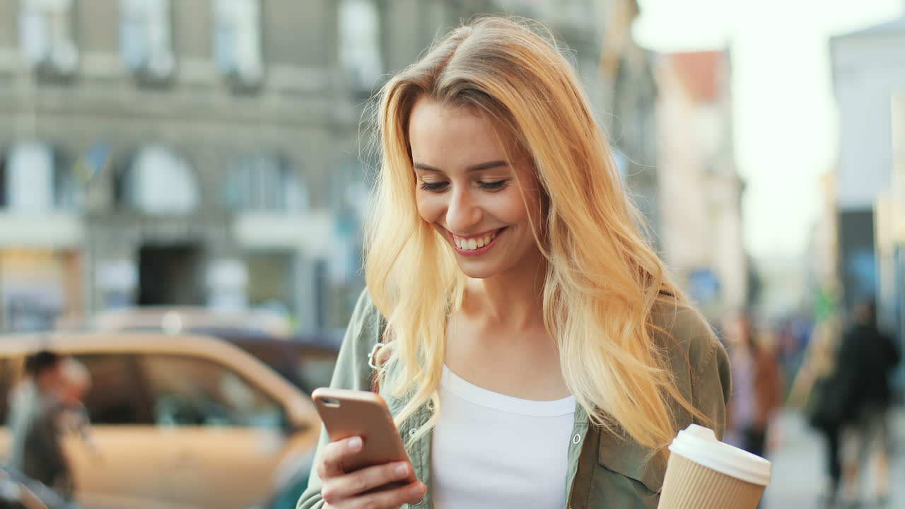 Close-up view of Caucasian blonde woman texting on the smartphone while walking down the street and holding coffee to go